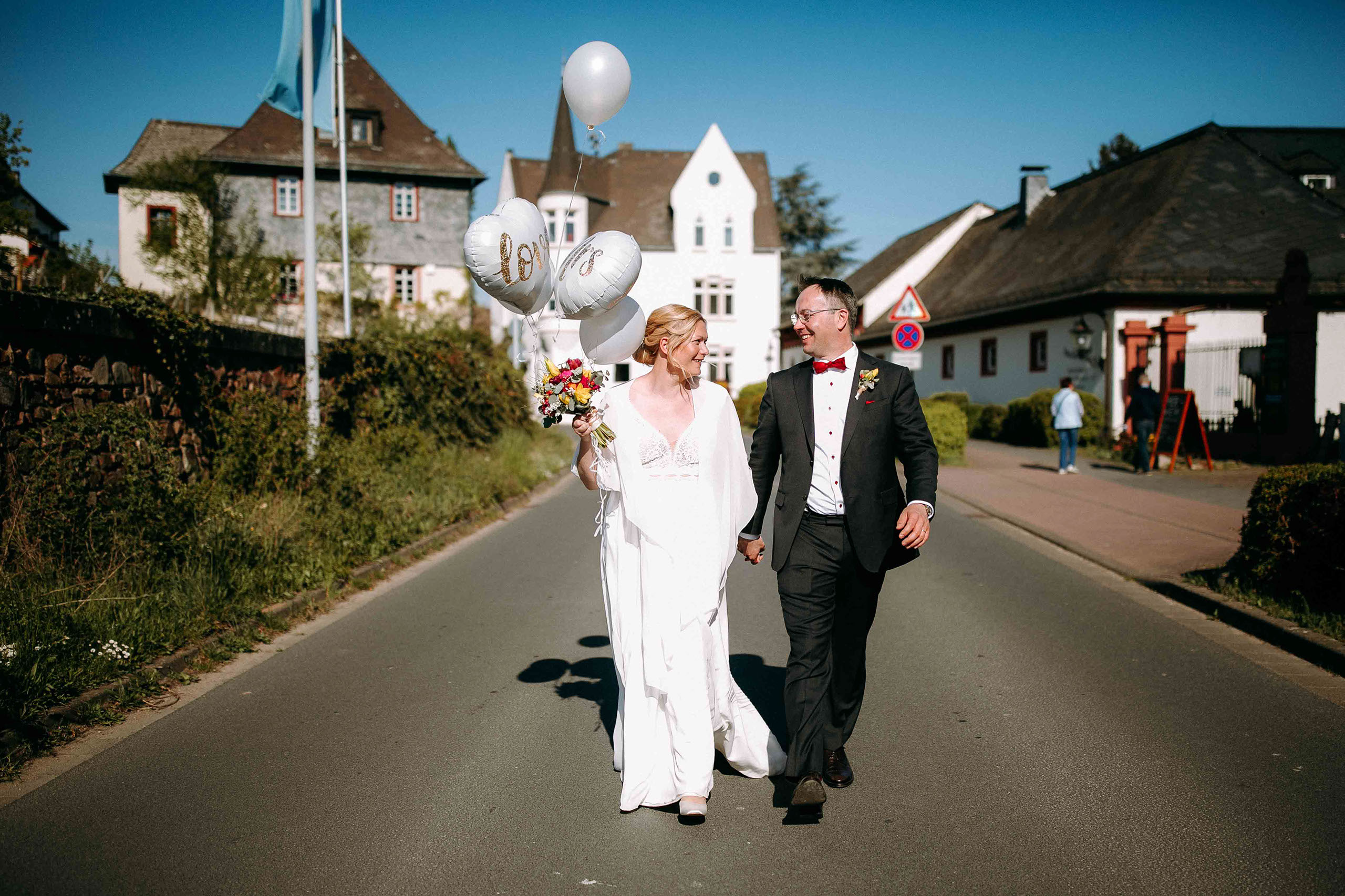 Fröhliches Brautpaar mit Herzballons auf der Straße vor Schloss Reinhartshausen – Hochzeitsreportage Rheingau