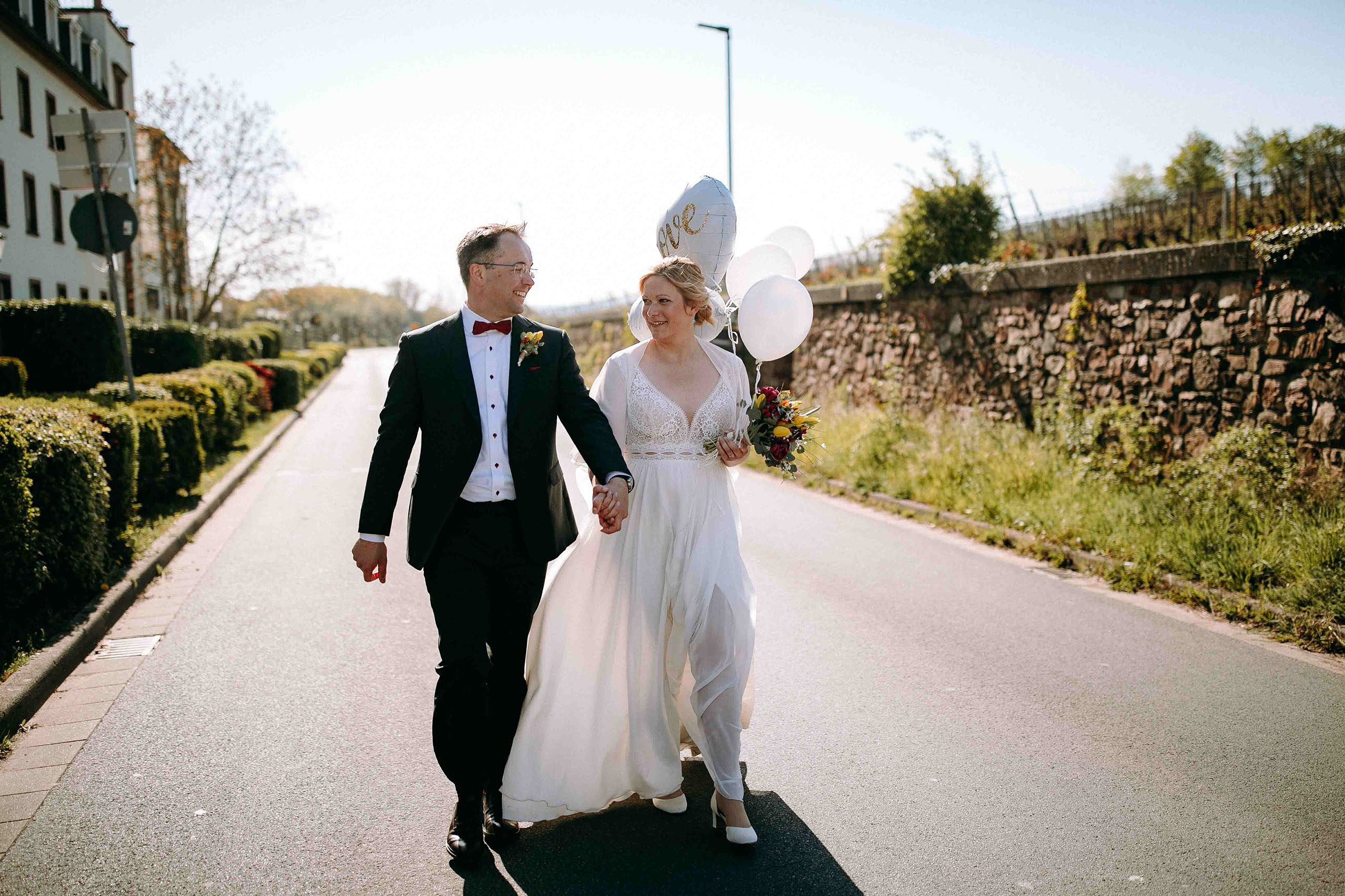 Braut und Bräutigam Hand in Hand auf Straße mit Blick auf Schloss und Weinberge – Hochzeitsfotograf Rheingau