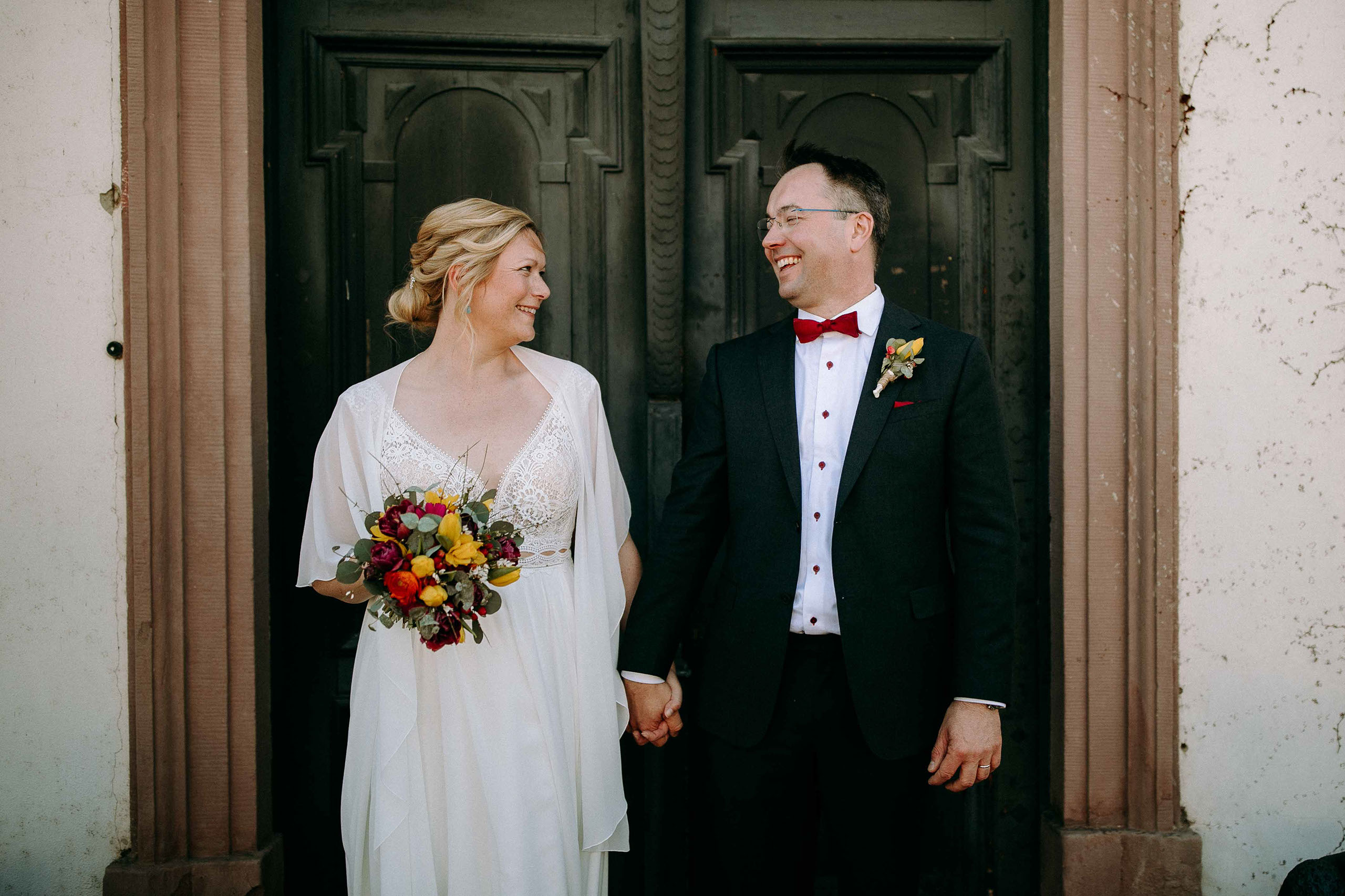 Braut und Bräutigam stehen vor antiker Tür und lachen sich an – Hochzeitslocation Schloss Reinhartshausen