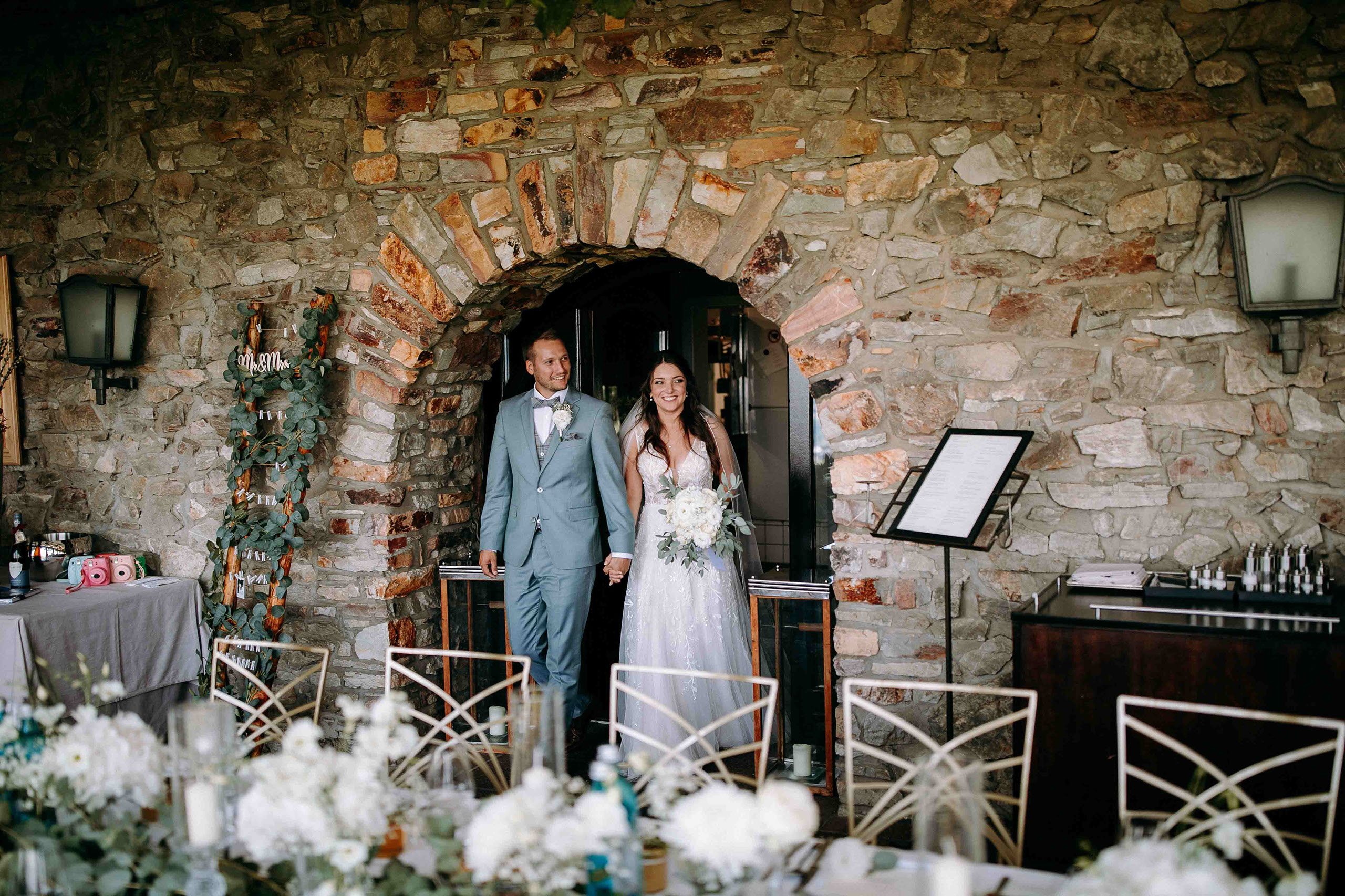Das Brautpaar betritt Händchen haltend die Hochzeitslocation Burg Schwarzenstein durch einen steinernen Torbogen – Hochzeitsfotograf Rheingau.