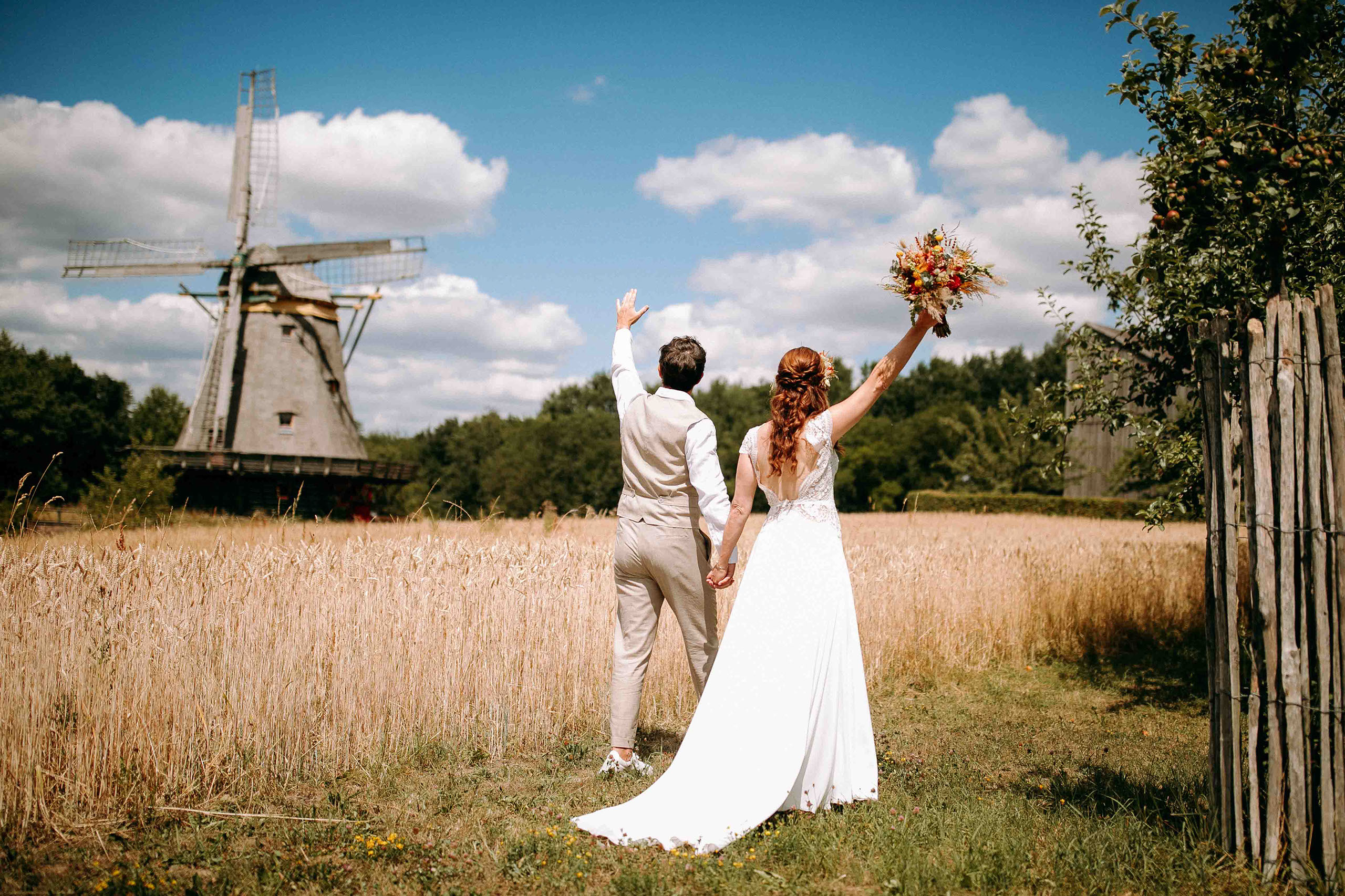 Braut und Bräutigam heben die Hände vor Freude vor einer Windmühle in die Luft.