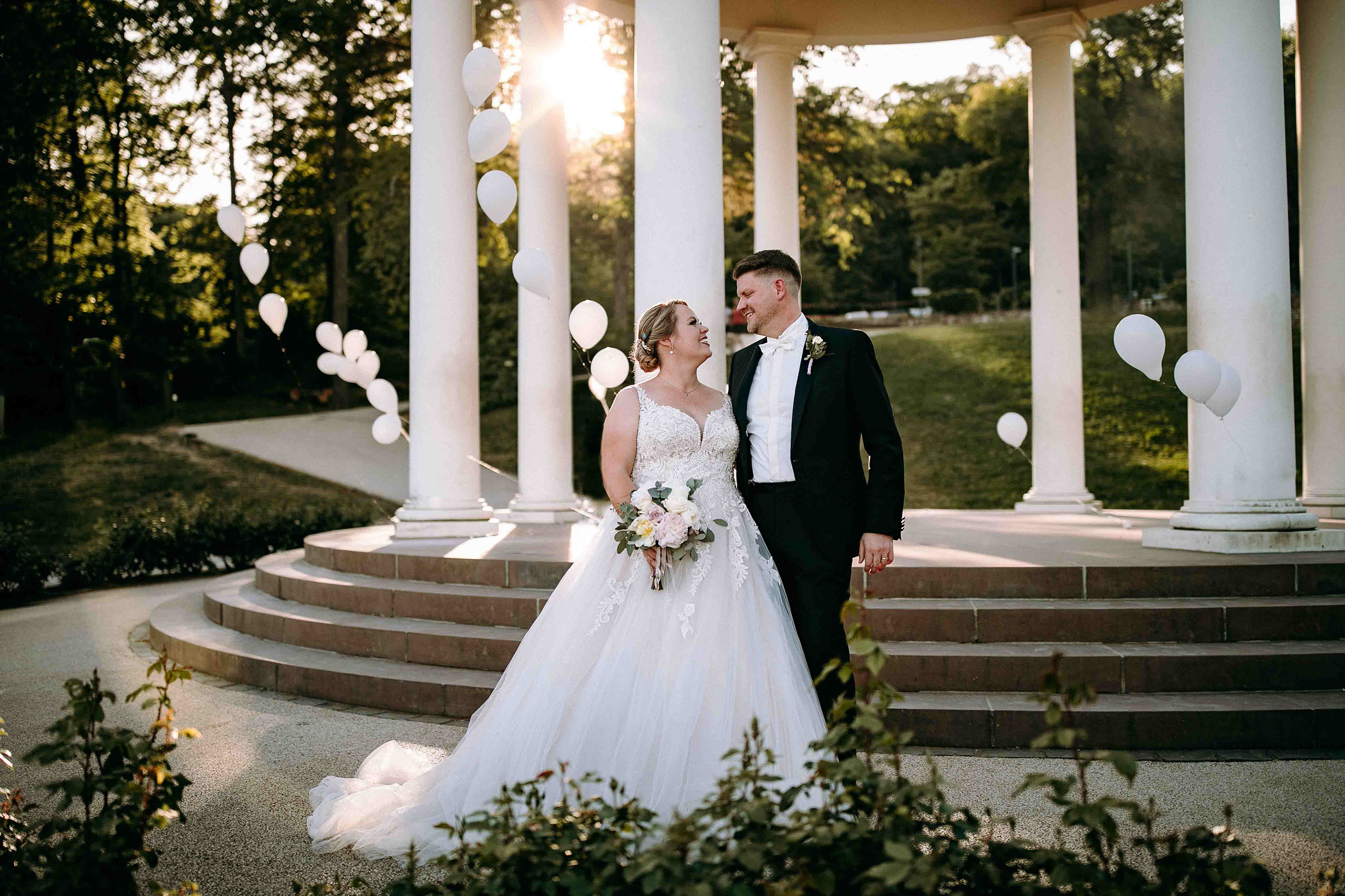 Brautpaar steht vor dem Niederwaldtempel mit weißen Ballons bei Sonnenuntergang – Hochzeitslocation Am Niederwald im Rheingau.