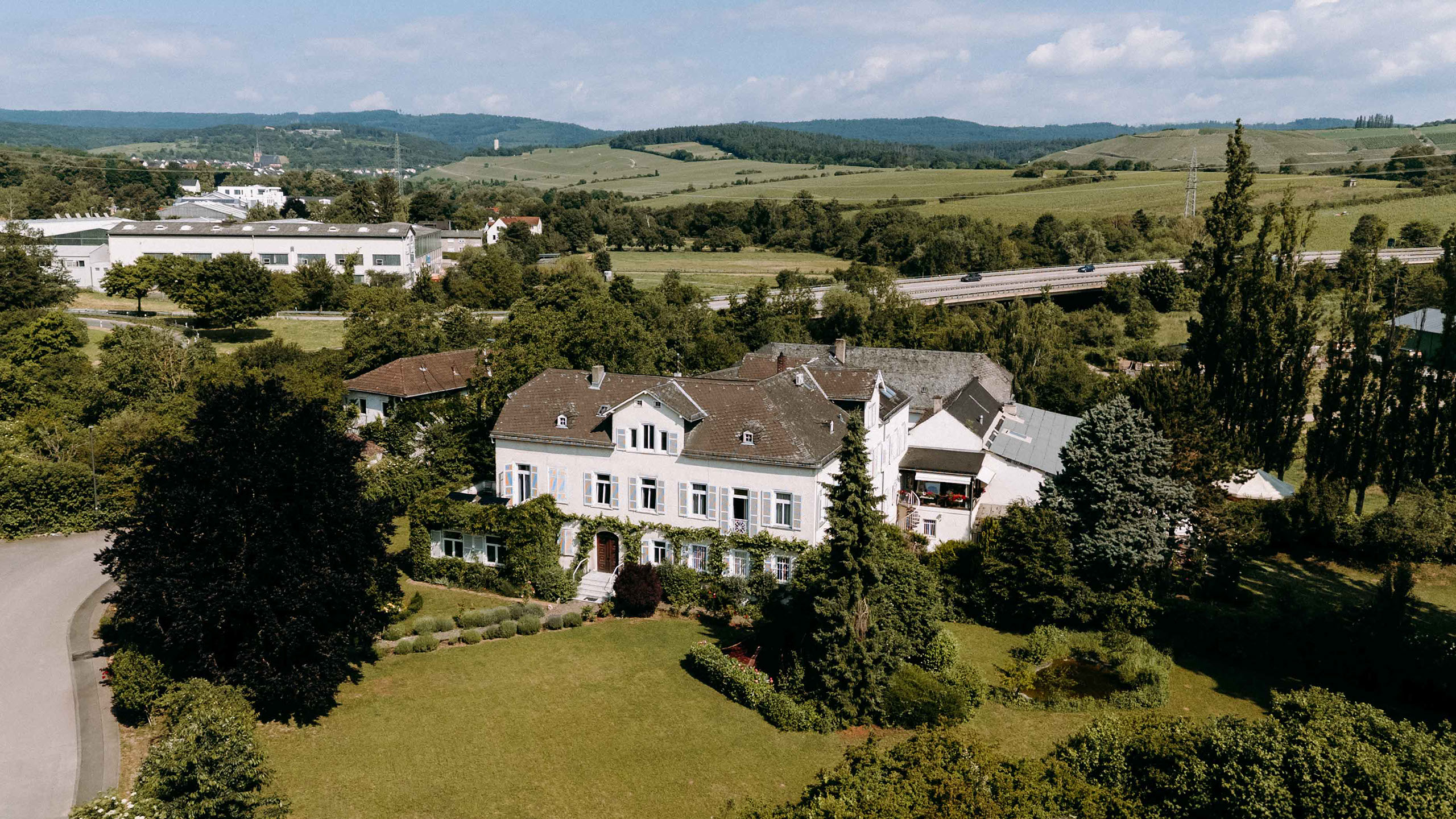 Drohnenansicht auf das elegante weiße Herrenhaus der Klostermuehle mit großem Garten,umgeben von Wald und Weinbergen. Hochzeitslocation Rheingau